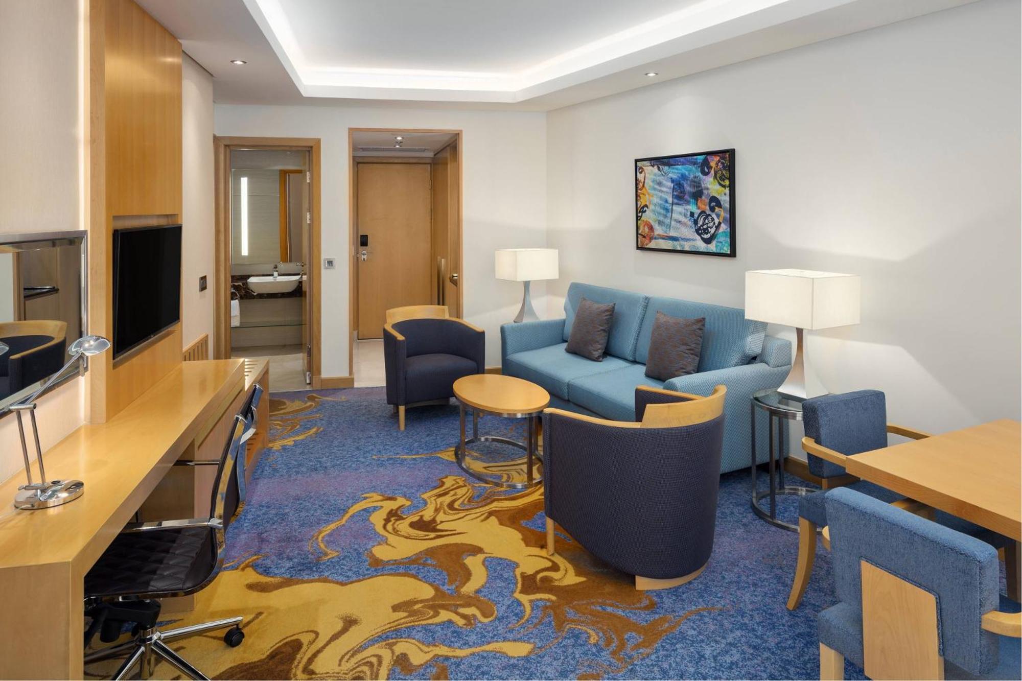 Sheraton Makkah Jabal Al Kaaba Hotel La Meca Exterior foto The photo displays a modern hotel room or suite. The room features a comfortable seating area with a blue couch and two blue armchairs surrounding a small round table. A wooden desk with a black office chair is positioned against one wall, accompanie