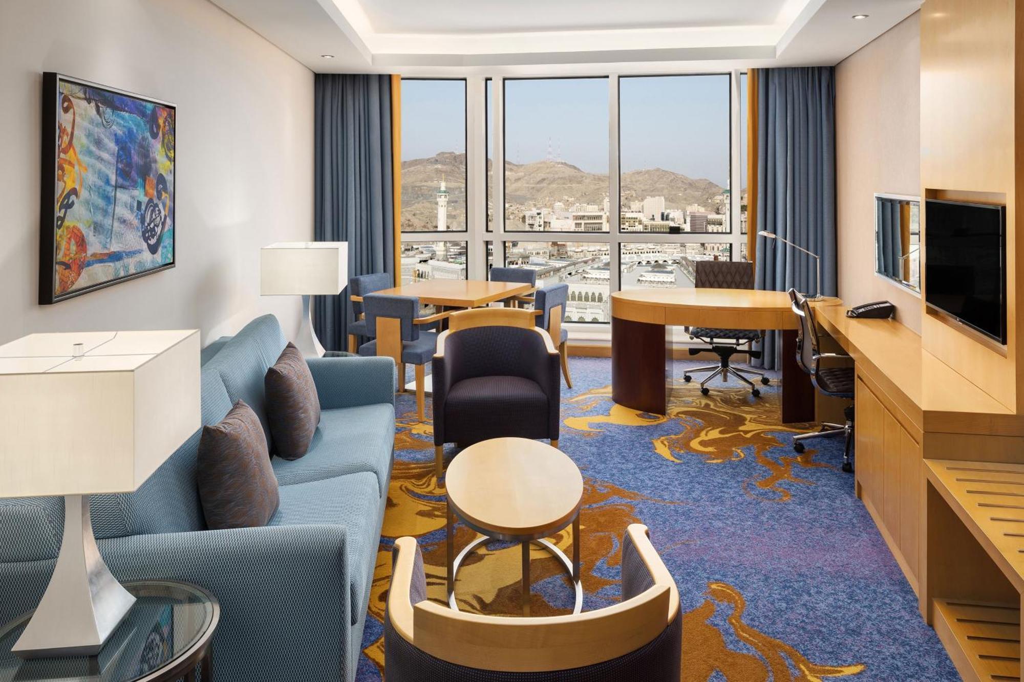 Sheraton Makkah Jabal Al Kaaba Hotel La Meca Exterior foto The photo shows a well-decorated hotel room featuring a modern and comfortable interior. There is a cozy seating area with a blue sofa and armchairs, complemented by a round coffee table. A desk with a chair is also present, ideal for work. The room 