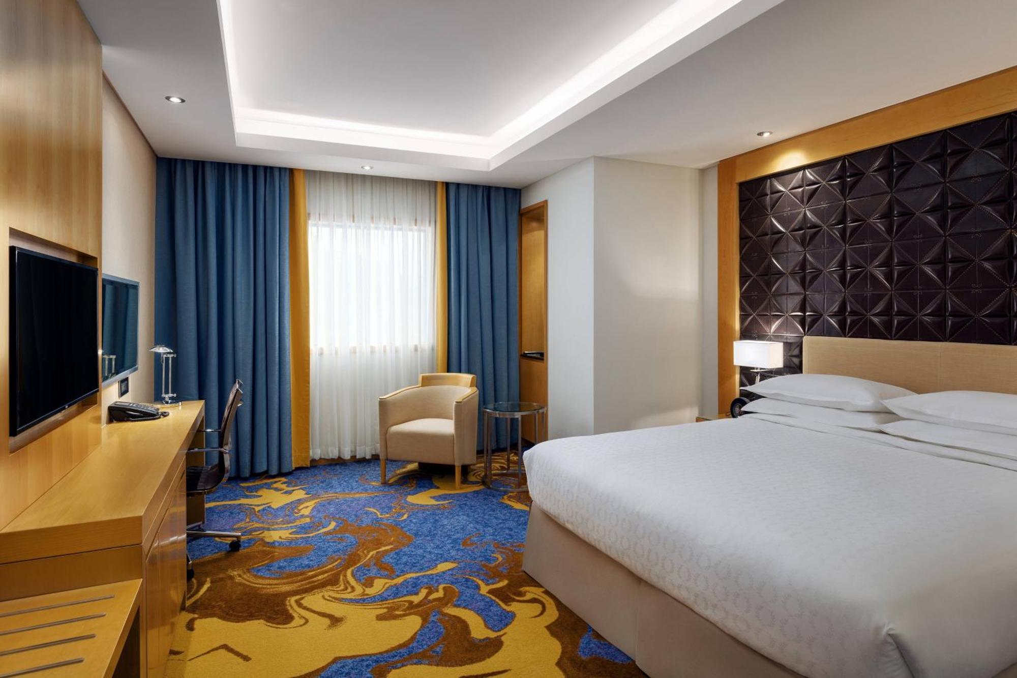 Sheraton Makkah Jabal Al Kaaba Hotel La Meca Exterior foto The photo shows a modern hotel room featuring a comfortable double bed with a light-colored headboard. There's a large window dressed with blue curtains, allowing natural light to enter. The walls are painted in neutral tones, and one wall has a text