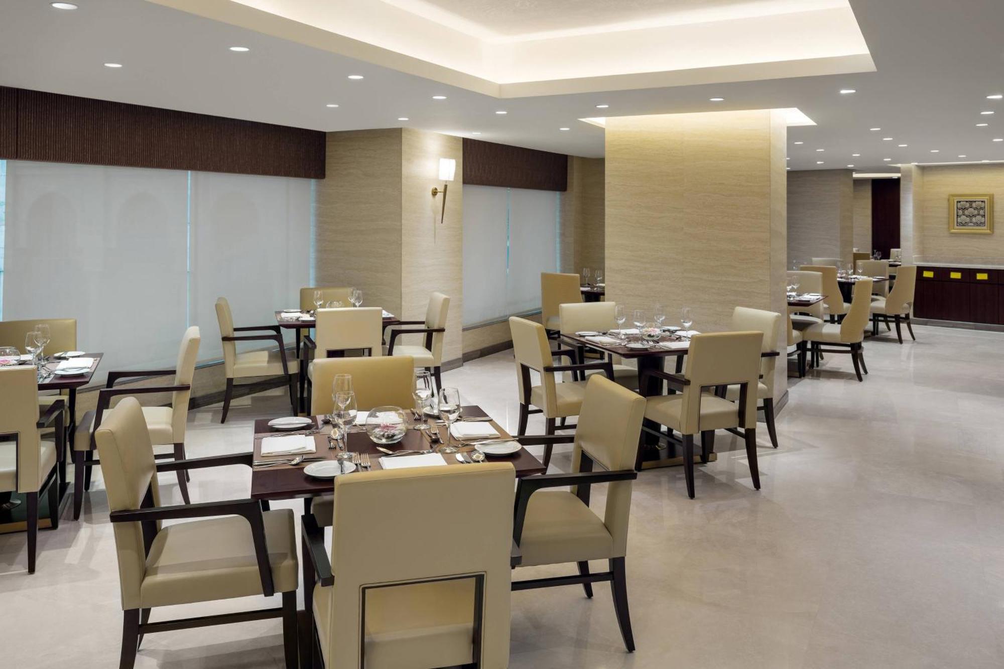 Sheraton Makkah Jabal Al Kaaba Hotel La Meca Exterior foto The photo shows a modern restaurant interior. It features several dining tables, each set with utensils and tableware. The chairs are upholstered in a light fabric, and the overall decor is sleek and minimalistic, with soft lighting from overhead fix